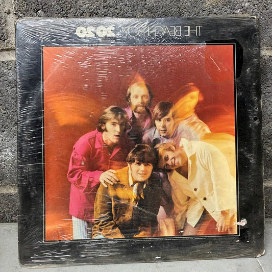 A vintage vinyl record album for The Beatles' '3 and 7 in the Morning' on the black sleeve, still sealed in its plastic shrink wrap, leaning against a gray concrete wall.