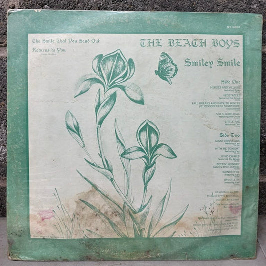 Vintage Beach Boys 'Smiley Smile' vinyl record album cover, featuring a green border and botanical illustration of iris flowers on an off-white background, leaning against a dark textured wall.
