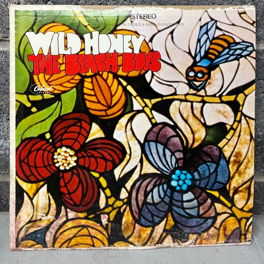 Vintage 'Wild Honey' album cover by The Beach Boys, featuring a stained-glass style floral design with a bee, photographed leaning against a gray concrete wall.