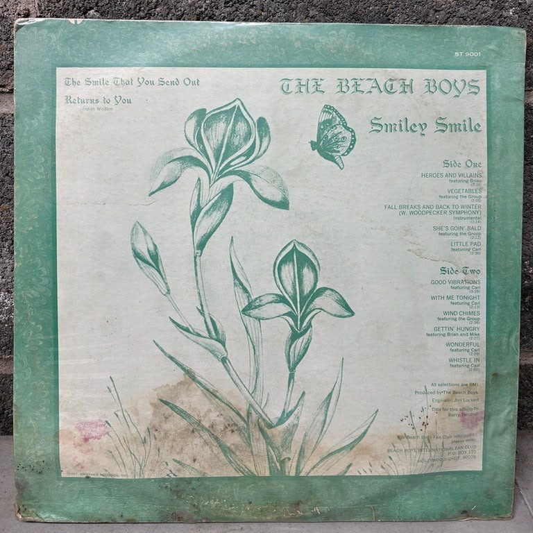 Vintage Beach Boys 'Smiley Smile' vinyl record album cover, featuring a green border and botanical illustration of iris flowers on an off-white background, leaning against a dark textured wall.