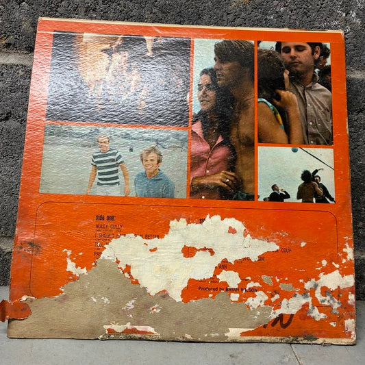 A square orange printed collage board or plaque with multiple small photos and a heavily worn, peeling orange-and-white lower section, photographed on a gray concrete surface.