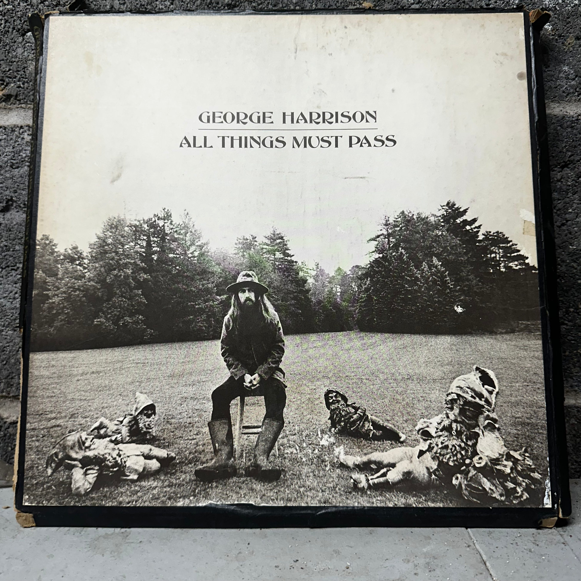 Album cover of 'All Things Must Pass' by George Harrison with a black and white photo of a man sitting on a stool with three people lying on the grass.