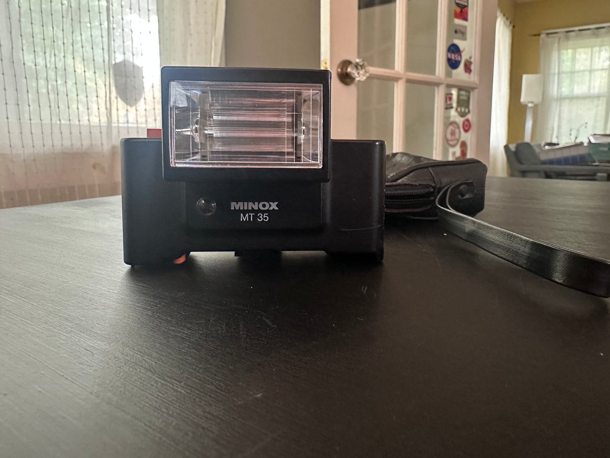 A Minox MT 35 film camera flash unit sitting on a dark wooden table, with its black strap coiled to the right and a blurred indoor room in the background.