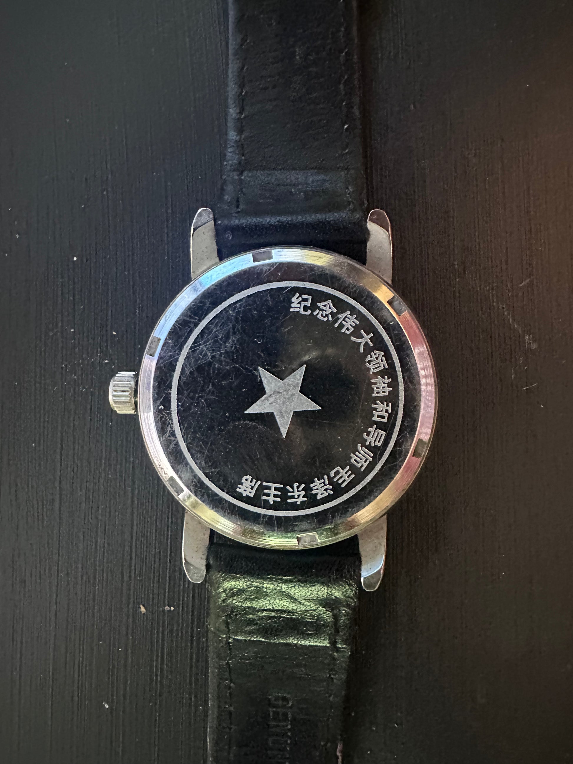 A vintage-style wristwatch with a round silver-tone case, black dial featuring a central star and Chinese text, and a black leather strap, shown on a dark textured background.