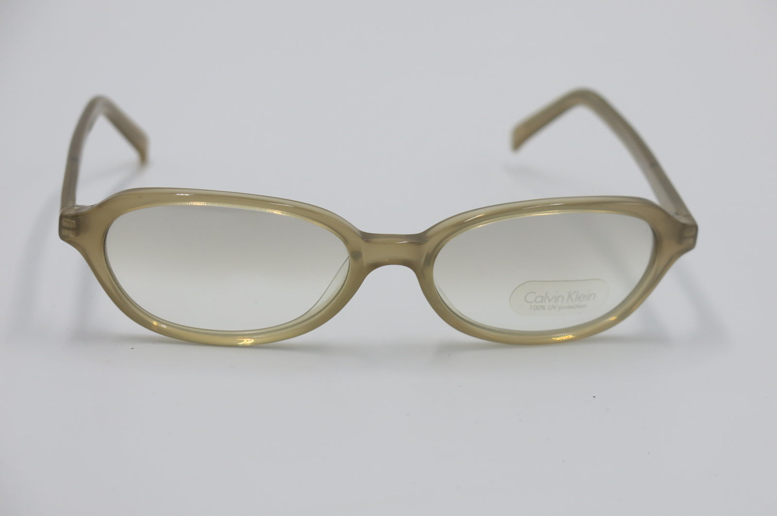 Oval eyeglasses with translucent olive-brown frames and clear lenses, featuring a visible Calvin Klein logo on the lower right lens, set against a plain light gray background.