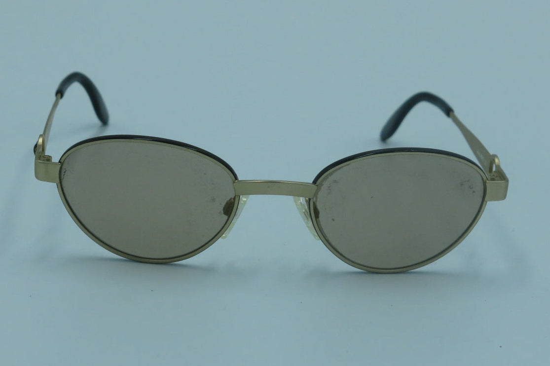 Oval sunglasses with a gold-tone metal frame and tinted gray lenses, shown on a light blue background.