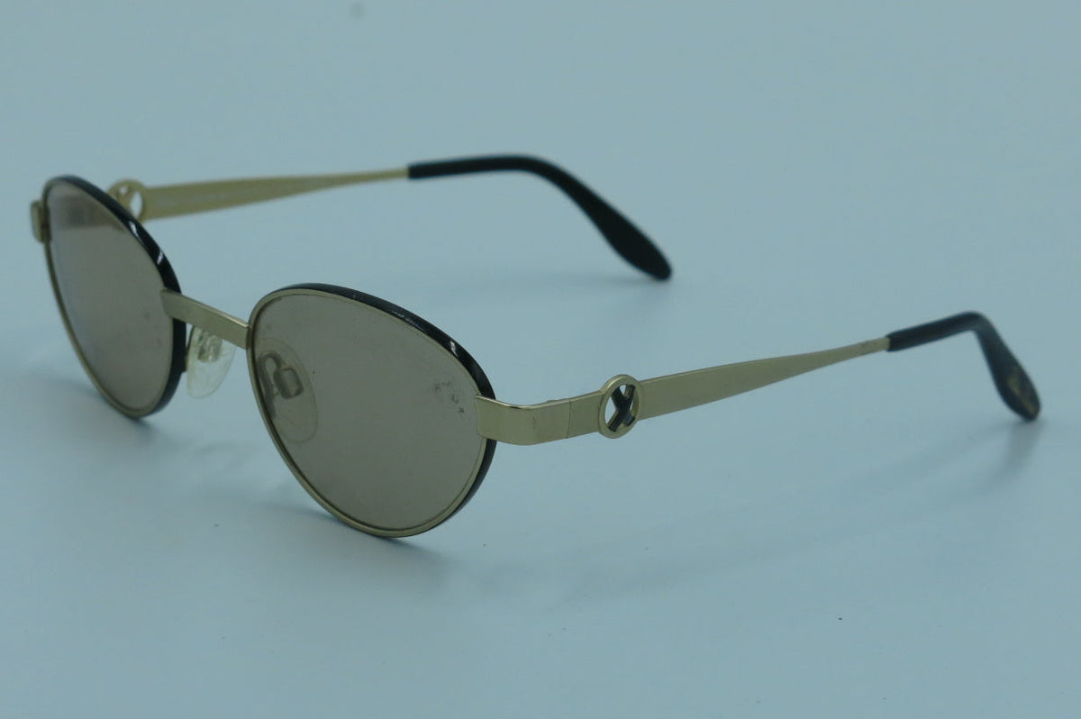 Oval sunglasses with gold-tone metal frames, dark lenses, and black temple tips, photographed on a light blue background.