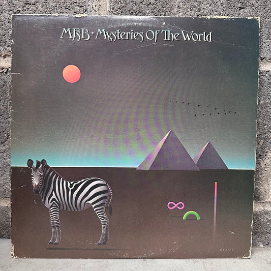 MFSB 'Mysteries Of The World' vinyl record album cover, featuring a zebra, pyramids, and a retro sci-fi landscape, photographed standing against a gray brick wall.