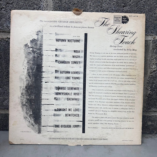 The back cover of a vintage vinyl record for George Shearing's 'The Shearing Touch' album, featuring a tracklist and track descriptions on an aged off-white background with black text and graphics, set against a dark gray textured wall.