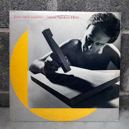 Vinyl record album cover for 'Music Spoken Here' by John McLaughlin, featuring a black and white photo of a shirtless man holding a wooden mallet over a board, with a large yellow abstract shape on a white background. The album is photographed standing against a gray textured wall.