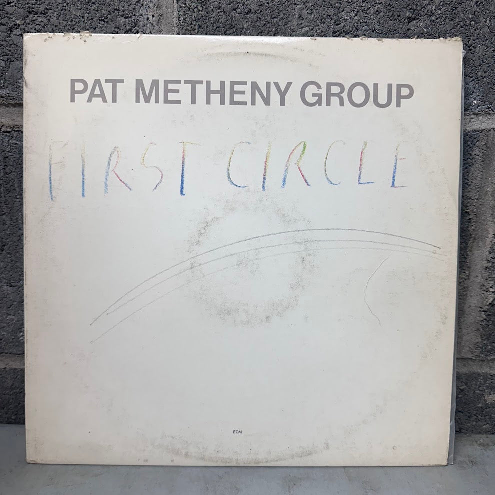 A vinyl record sleeve for 'First Circle' by the Pat Metheny Group, photographed standing against a dark gray brick wall. The sleeve is off-white with the artist's name printed in dark gray at the top and the album title written in a multicolored, handwritten style across the center.