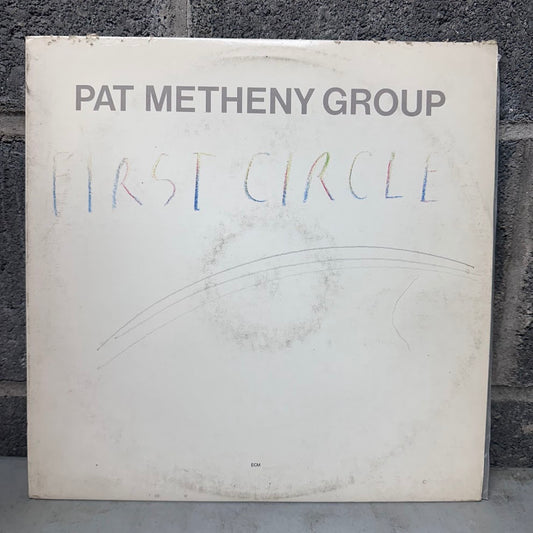 A vinyl record sleeve for 'First Circle' by the Pat Metheny Group, photographed standing against a dark gray brick wall. The sleeve is off-white with the artist's name printed in dark gray at the top and the album title written in a multicolored, handwritten style across the center.