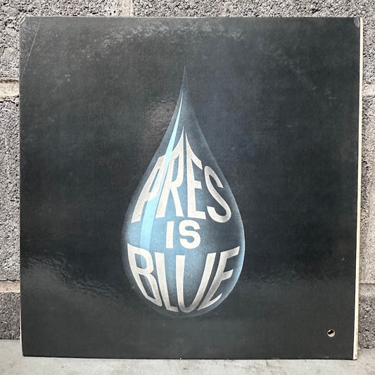 A square vinyl record sleeve or album cover leaning against a gray brick wall. The cover is predominantly black with a large glossy teardrop graphic in the center containing the words 'PRES IS BLUE' in white and light blue.